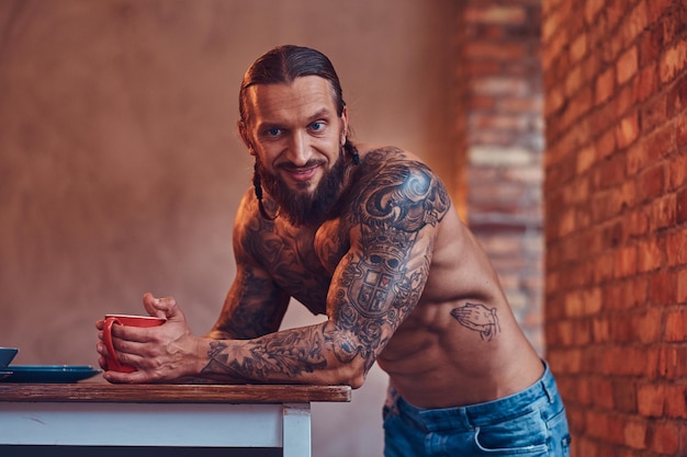 Bel maschio tatuato barbuto con un taglio di capelli elegante e un corpo muscoloso, beve caffè, appoggiandosi a un tavolo.