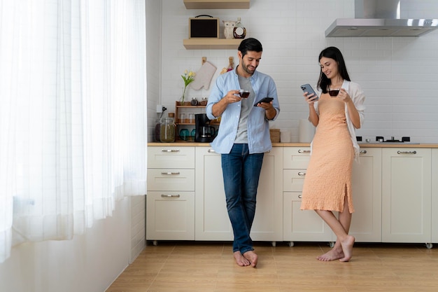 Handsome bearded young man and beautiful young woman standing in kitchen drinking morning coffee using mobile smartphone