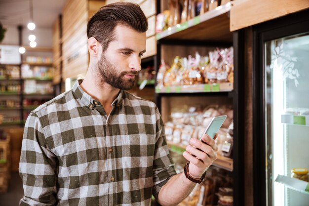 Bel giovane barbuto in camicia a quadri usando il cellulare nel negozio di alimentari