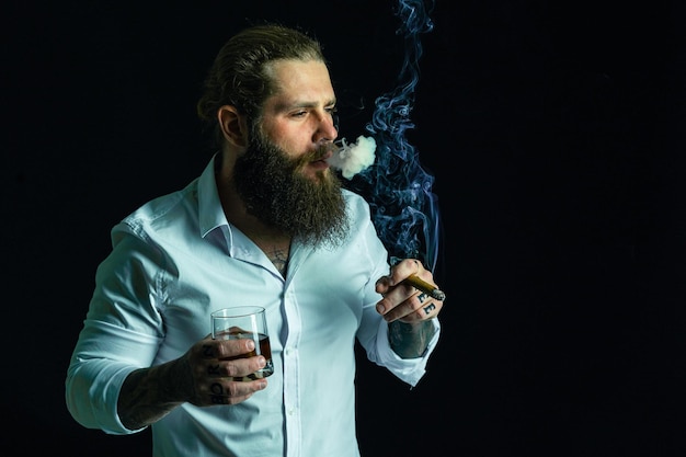 Красивый бородатый молодой человек курит сигару, держит стакан виски, одетый в белую рубашку, студийный снимок
