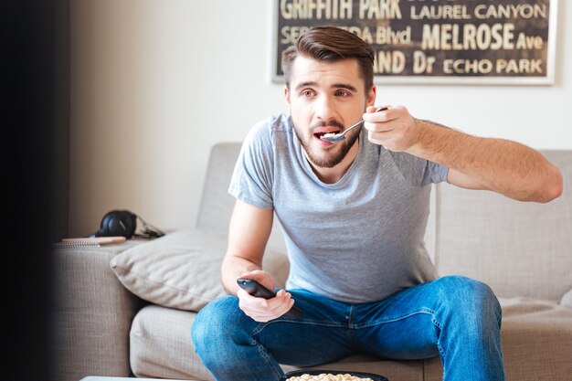 Handsome bearded young man with remote control sitting on sofa and eating sereals