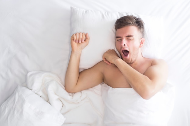 Handsome beautiful nice guy, young sleepy man yawning with open mouth in bed on pillow, covered with white blanket, ready for sleep or just wake up. Healthy good sleeping at home in bedroom. Top view