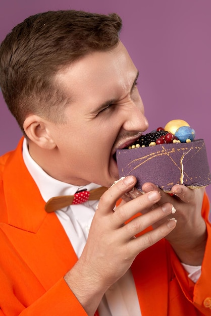 Photo handsome birthday man in orange jacket bites cake on purple background