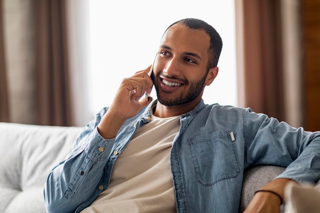 Handsome Black Guy Talking On Cellphone While Sitting On Couch At Home