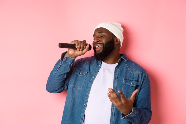 Handsome Black man in beanie and denim shirt singing karaoke, holding microphone, standing over pink background