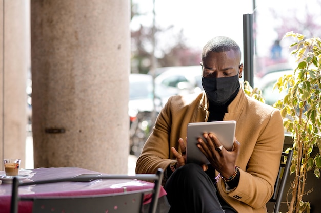 Handsome black man in elegant stylish suit sitting in a coffee table using tablet. Guy wearing face mask.