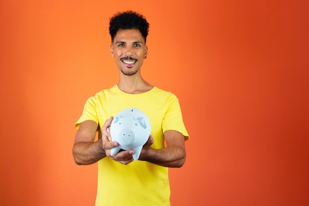 Uomo nero bello che tiene un salvadanaio isolato su orange uomo in una camicia gialla isolata