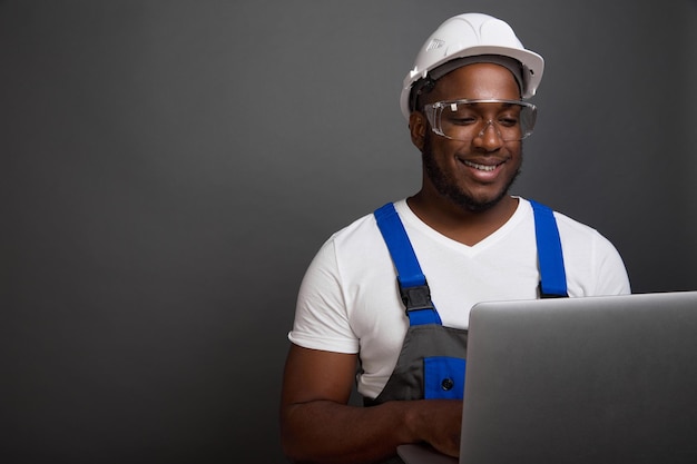 Handsome black man in white helmet and overalls with a smile works at a laptop