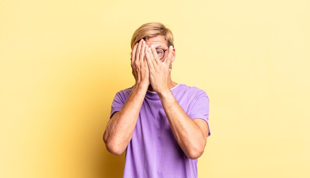 Handsome blond adult man covering face with hands, peeking between fingers with surprised expression and looking to the side