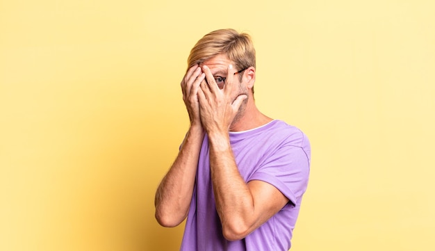 Handsome blond adult man feeling scared or embarrassed, peeking or spying with eyes half-covered with hands