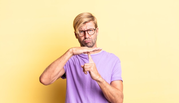 Handsome blond adult man looking serious, stern, angry and displeased, making time out sign