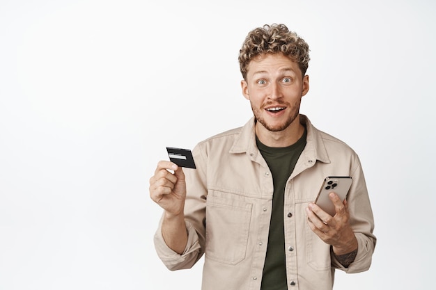 Handsome blond curly guy holding smartphone and credit card looking excited about store sale paying online order in mobile phone white background