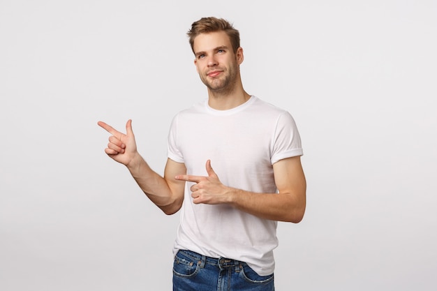 Handsome blond guy with blue eyes and white T-shirt pointing to the side