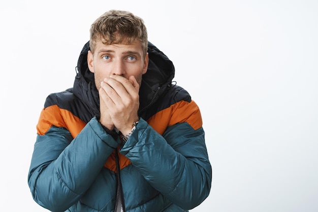 Handsome blond man with blue eyes exhaling air in palms to warmup standing in stylish puffer jacket outside