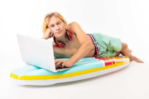 handsome blond man with laptop and headphones sits on an air mattress for swimming on a white  wall