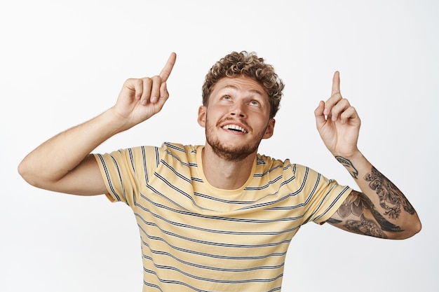 Handsome blond smiling man pointing fingers up bending while looking up with excited face expression standing in casual clothes over white background