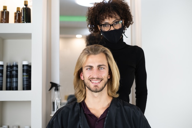 黒人女性の美容師によって散髪されているハンサムなブロンドの男性のクライアント