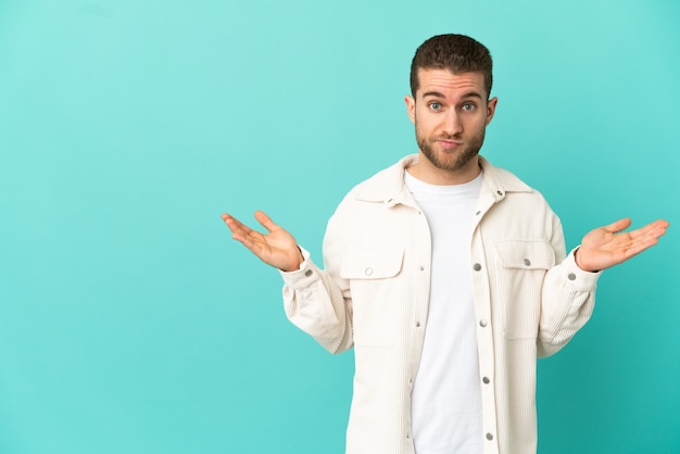 Handsome blonde man over isolated blue background having doubts while raising hands