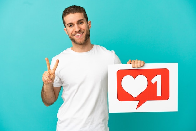 Handsome blonde man over isolated blue background holding a placard with Like icon and celebrating a victory