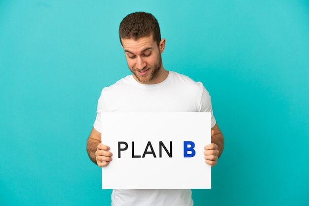 Handsome blonde man over isolated blue background holding a placard with the message PLAN B