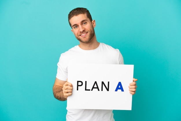 Handsome blonde man over isolated blue background holding a placard with the message PLAN A