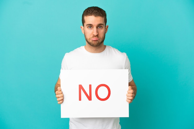 Handsome blonde man over isolated blue background holding a placard with text NO with sad expression