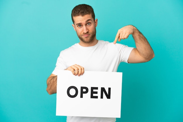 Handsome blonde man over isolated blue background holding a placard with text OPEN and  pointing it