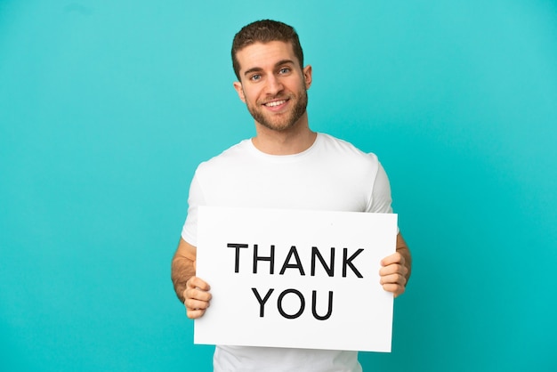 Handsome blonde man over isolated blue background holding a placard with text THANK YOU with happy expression