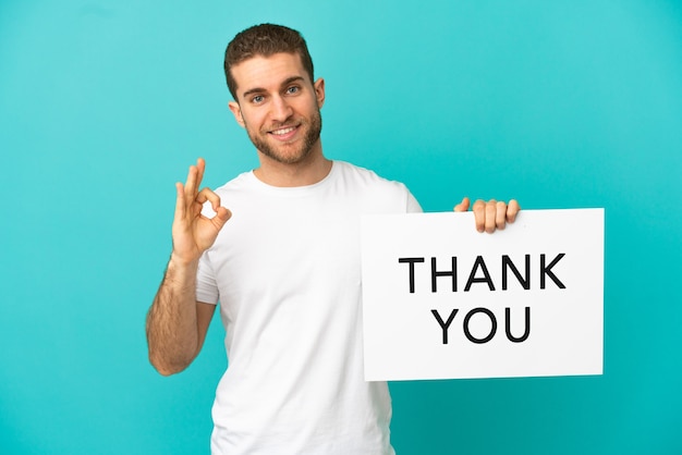 Handsome blonde man over isolated blue background holding a placard with text THANK YOU with ok sign