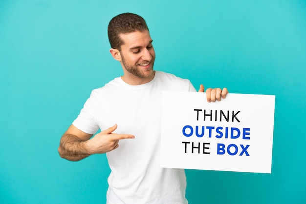 Handsome blonde man over isolated blue background holding a placard with text Think Outside The Box and  pointing it