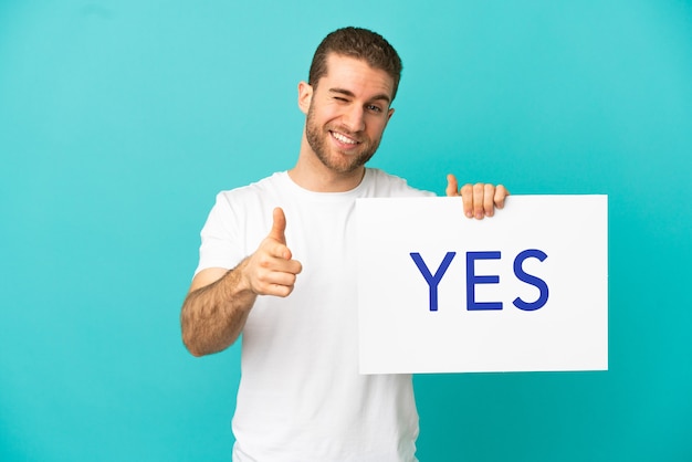 Handsome blonde man over isolated blue background holding a placard with text YES and pointing to the front