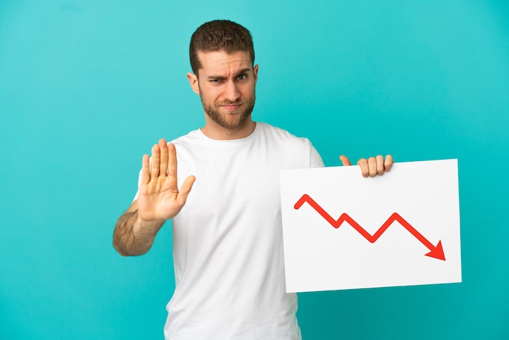 फोटो handsome blonde man over isolated blue background holding a sign with a decreasing statistics arrow symbol and doing stop sign