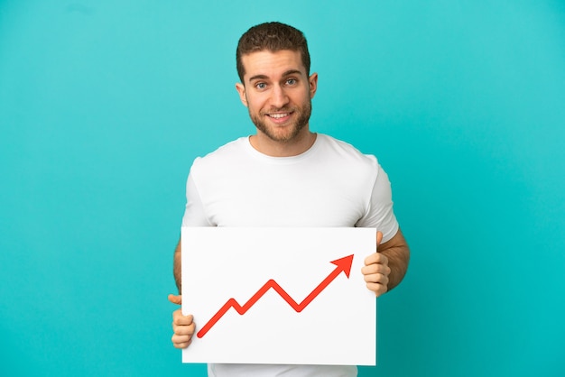 Handsome blonde man over isolated blue background holding a sign with a growing statistics arrow symbol with happy expression