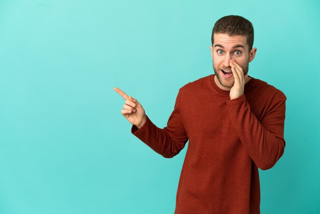 Handsome blonde man over isolated blue background pointing to the side to present a product and whispering something