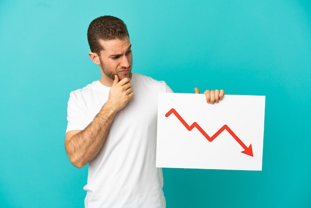 Handsome blonde man over isolated blue wall holding a sign with a decreasing statistics arrow symbol and thinking