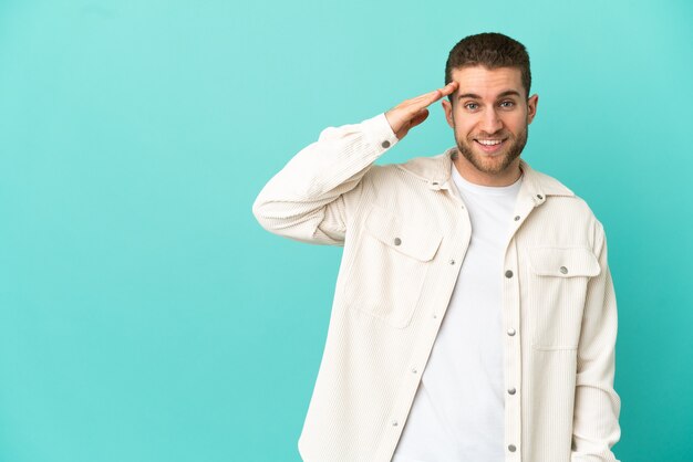 Handsome blonde man over isolated blue wall saluting with hand with happy expression