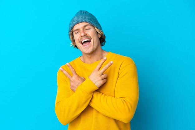 Handsome blonde man isolated on blue wall smiling and showing victory sign