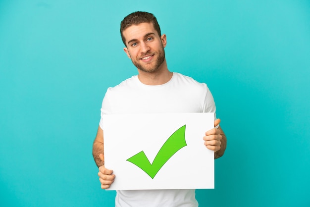 Handsome blonde man over isolated holding a placard with text Green check mark icon with happy expression