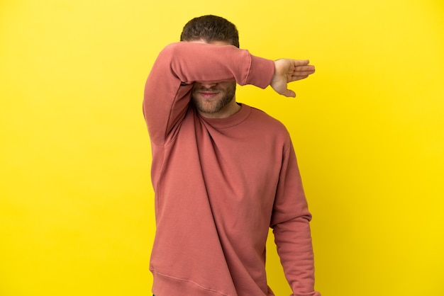 Handsome blonde man over isolated yellow background covering eyes by hands