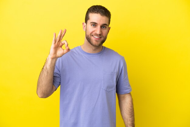 Handsome blonde man over isolated yellow background showing ok sign with fingers