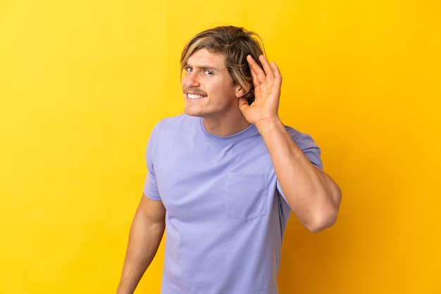 Photo handsome blonde man isolated on yellow listening to something by putting hand on the ear