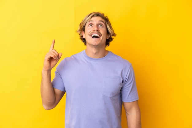 Handsome blonde man isolated on yellow pointing up and surprised