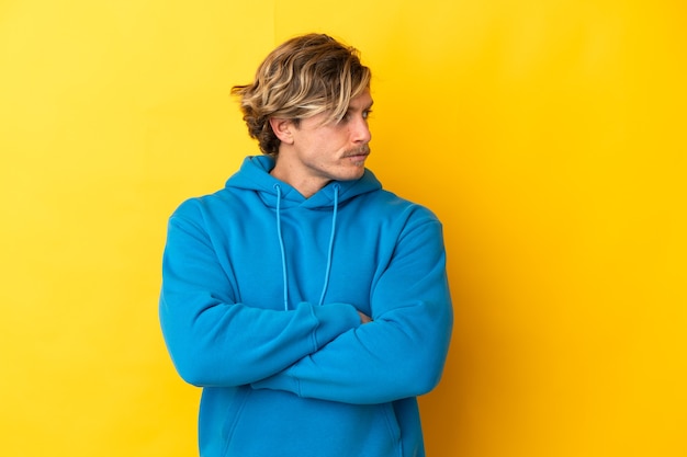 Handsome blonde man isolated on yellow wall keeping the arms crossed