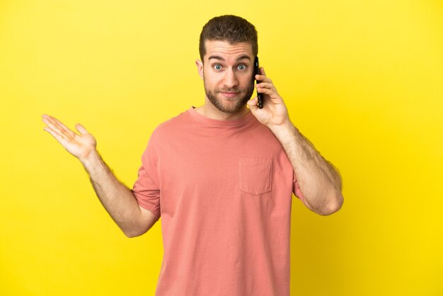 Handsome blonde man using mobile phone over isolated background having doubts while raising hands