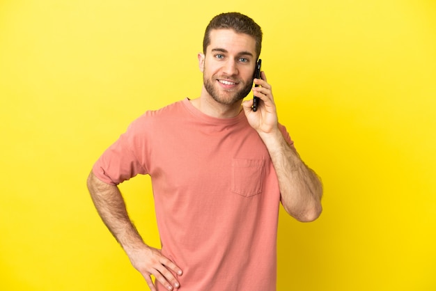 Handsome blonde man using mobile phone over isolated background posing with arms at hip and smiling