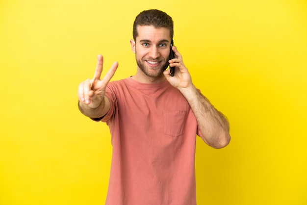 Bel uomo biondo che utilizza il telefono cellulare su sfondo isolato sorridente e mostrando segno di vittoria