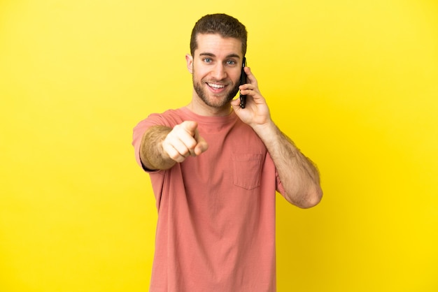 Handsome blonde man using mobile phone over isolated background surprised and pointing front