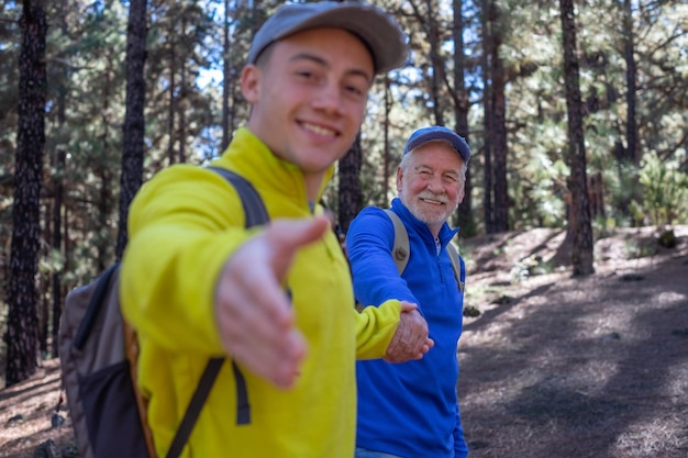Handsome blurred boy walks happily in the forest holding his\
grandfather\'s hand smiling multigenerational family enjoying\
freedom mountain and nature together the new generation helps the\
old one