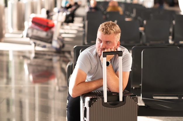 Handsome bored tired unhappy caucasian passenger man sitting\
with suitcase baggage at airport waiting for checkin\
registrationconcept of flight delay