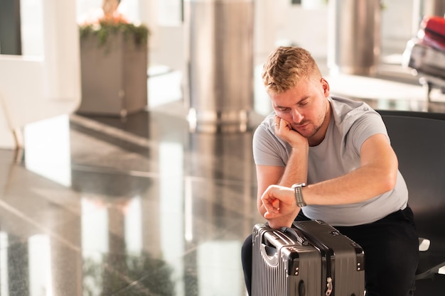 Handsome boring tired unhappy caucasian passenger man sitting with suitcase baggage at airport waiting for checkin registration looking at watchConcept of flight delay